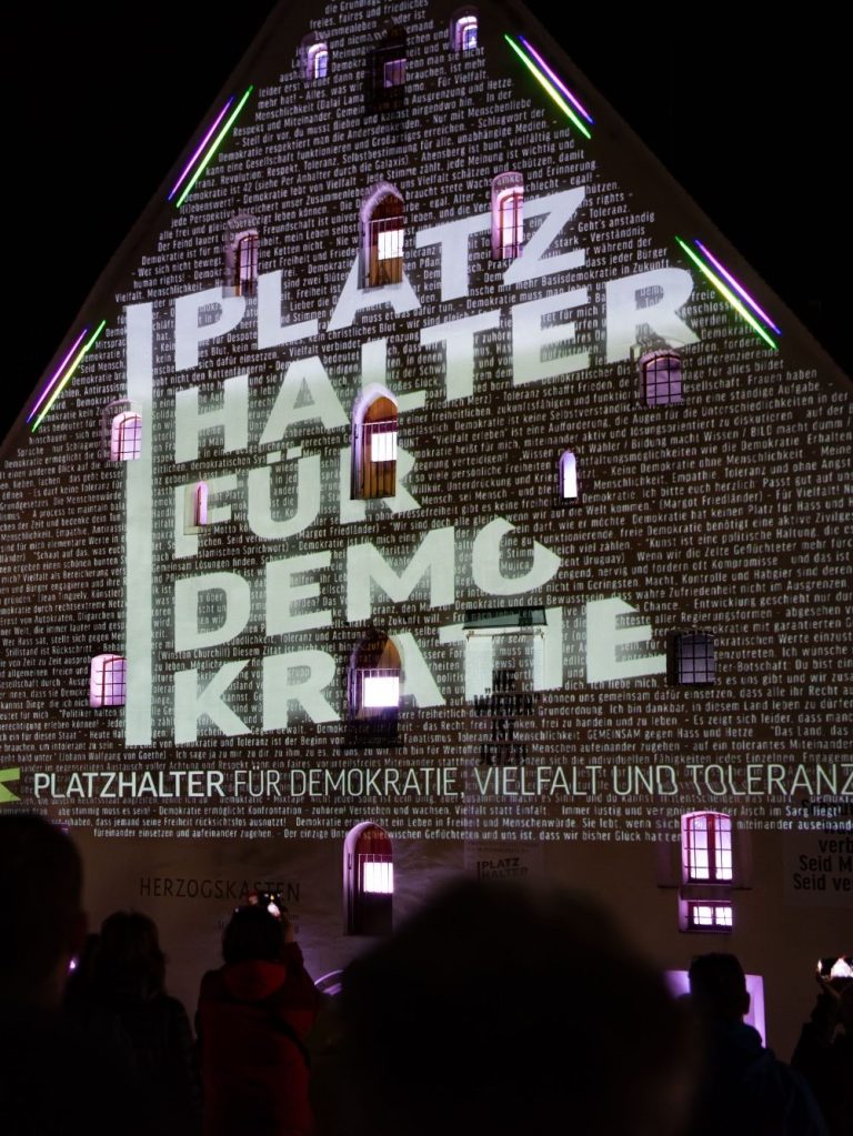 Platzhalter Logo Projektion Fassade des Herzogskastens mit Projektion ‚PLATZHALTER DEMOKRATIE‘ und ‚PLATZHALTER FÜR DEMOKRATIE, VIELFALT UND TOLERANZ‘.