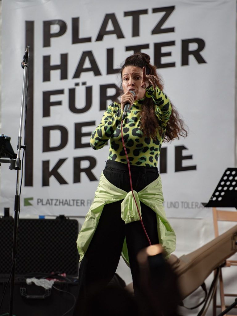 RiA REiSER Sängerin RiA REiSER mit Mikrofon auf der Bühne vor Banner ‚PLATZ HALTER FÜR DEMOKRATIE‘.