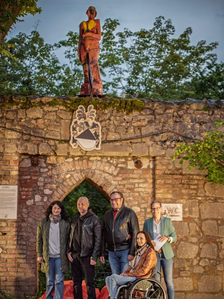 Vertreter des Platzhalter Kollektives, Bildhauer Klaus Schwendner, 1. Bürgermeister Dr. Bernhard Resch, 3. Bürgermeisterin Marion Huber-Schallner und Museumsleiterin Veronika Wimmer bei der Enthüllung der Skulptur "DAVINA" im Schlossgarten Fünf Personen vor alter Steinmauer mit Wappen und Holzskulptur ‚Davina‘ auf der Schlossgartenmauer.